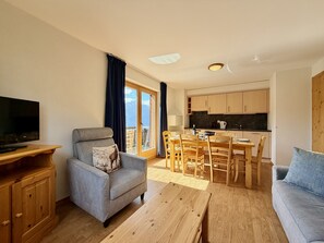 A modern living room with a sofa, a television, a dining table with chairs, and a kitchen area.