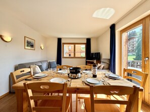 A dining area with a wooden table set for four, surrounded by chairs, with a television and a window in the background.