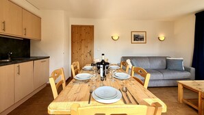 A dining area with a wooden table set for four, surrounded by wooden chairs. There is a kitchen area with cabinets and a countertop to the left. A grey sofa is against the wall, and two wall-mounted lights are visible. - Pracondu 1 405 - OUTDOOR & FUN  apartment 6 pers (Haute-Nendaz)