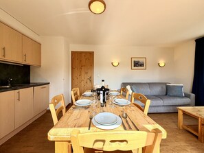 Ein Essbereich mit einem Holztisch, gedeckt für vier Personen, umgeben von Holzsesseln. Links befindet sich eine Küchenzeile mit Schränken und einer Arbeitsplatte. Eine graue Couch steht an der Wand, und zwei Wandleuchten sind sichtbar.
