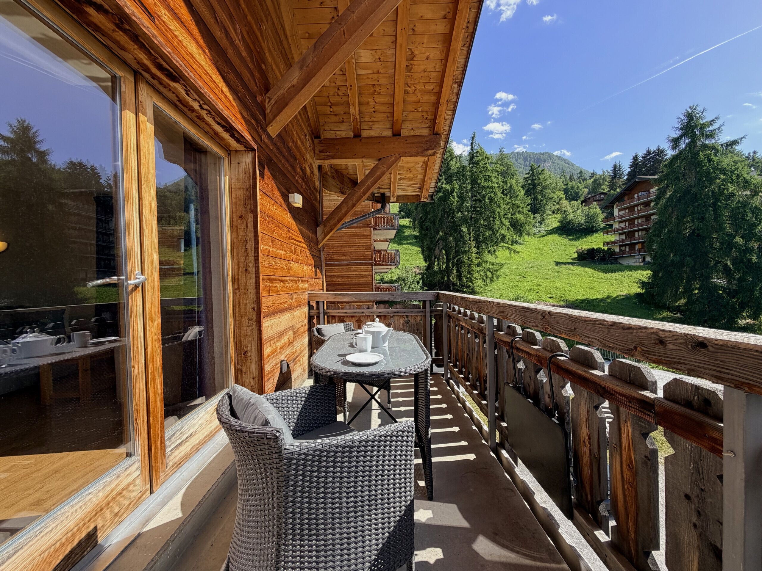 A wooden balcony with wicker furniture, a glass door, and a view of a green landscape with buildings in the background.