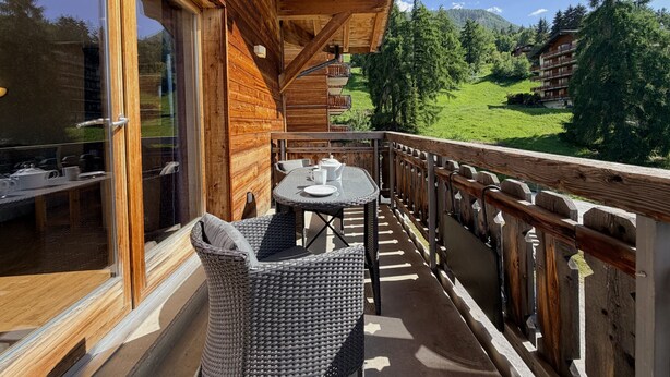 A wooden balcony with wicker furniture, a glass door, and a view of a green landscape with buildings in the background. - Pracondu 1 405 - OUTDOOR & FUN  apartment 6 pers (Haute-Nendaz)