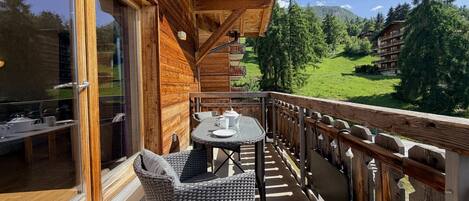 Un balcon en bois agrémenté de meubles en osier, une porte vitrée et une vue sur un paysage verdoyant, avec des bâtiments en arrière-plan.