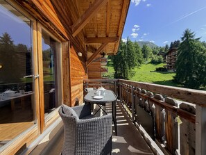 A wooden balcony with wicker furniture, a glass door, and a view of a green landscape with buildings in the background. - Pracondu 1 405 - OUTDOOR & FUN  apartment 6 pers (Haute-Nendaz)