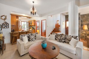 A living room with a sofa set, a coffee table, a chandelier, and a kitchen area visible through an open door.
