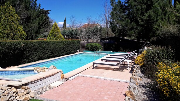 Une piscine rectangulaire dont les bords sont recouverts de carreaux, entourée d’un mur de pierre et d’une terrasse dallée. On aperçoit des chaises longues et une petite structure en bois au fond.