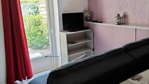 Une pièce comprenant un canapé, un téléviseur posé sur un meuble, une étagère avec des objets décoratifs et une fenêtre donnant sur un paysage verdoyant.