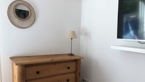 A wooden dresser with three drawers, a wall-mounted mirror, a lamp, and a TV on a white shelf.