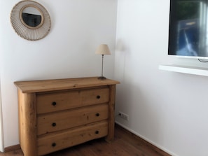 A wooden dresser with three drawers, a wall-mounted mirror, a lamp, and a TV on a white shelf.