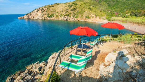 A coastal area with clear blue waters, rocky terrain, and a seating area with green and white striped lounge chairs and red umbrellas.