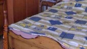 Ein Zimmer mit einem hölzernen Bett, einem Schrank aus Holz, einem kleinen Nachttisch mit Lampe und einem an der Wand befestigten Lichtschalter.