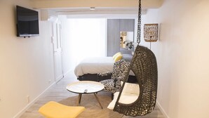 Une chambre d’hôtel moderne, équipée d’un lit, d’un fauteuil suspendu, d’une petite table et d’un téléviseur fixé au mur.