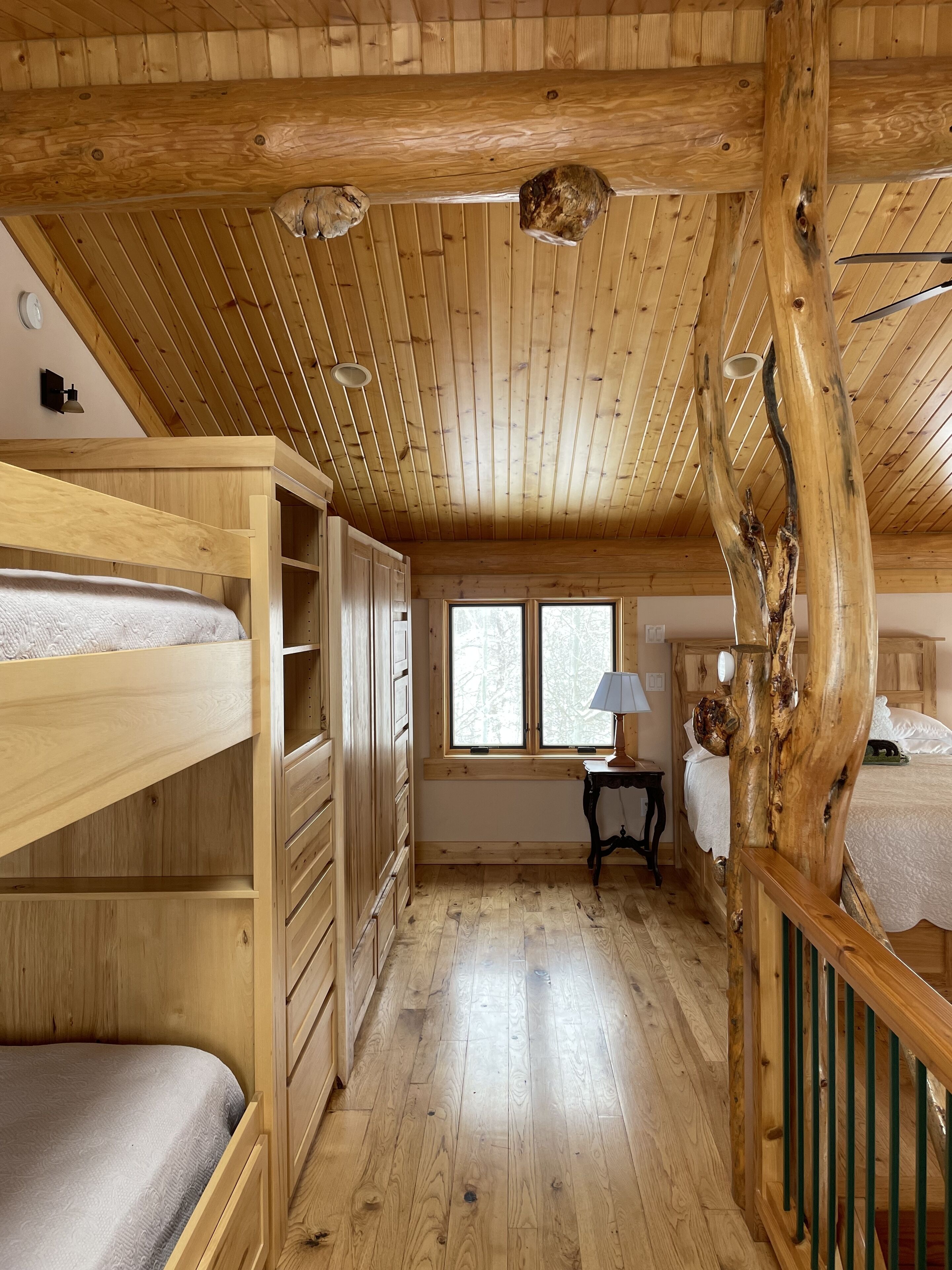 A cabin interior with wooden floors, bunk beds, a staircase, and a window with a view of trees.