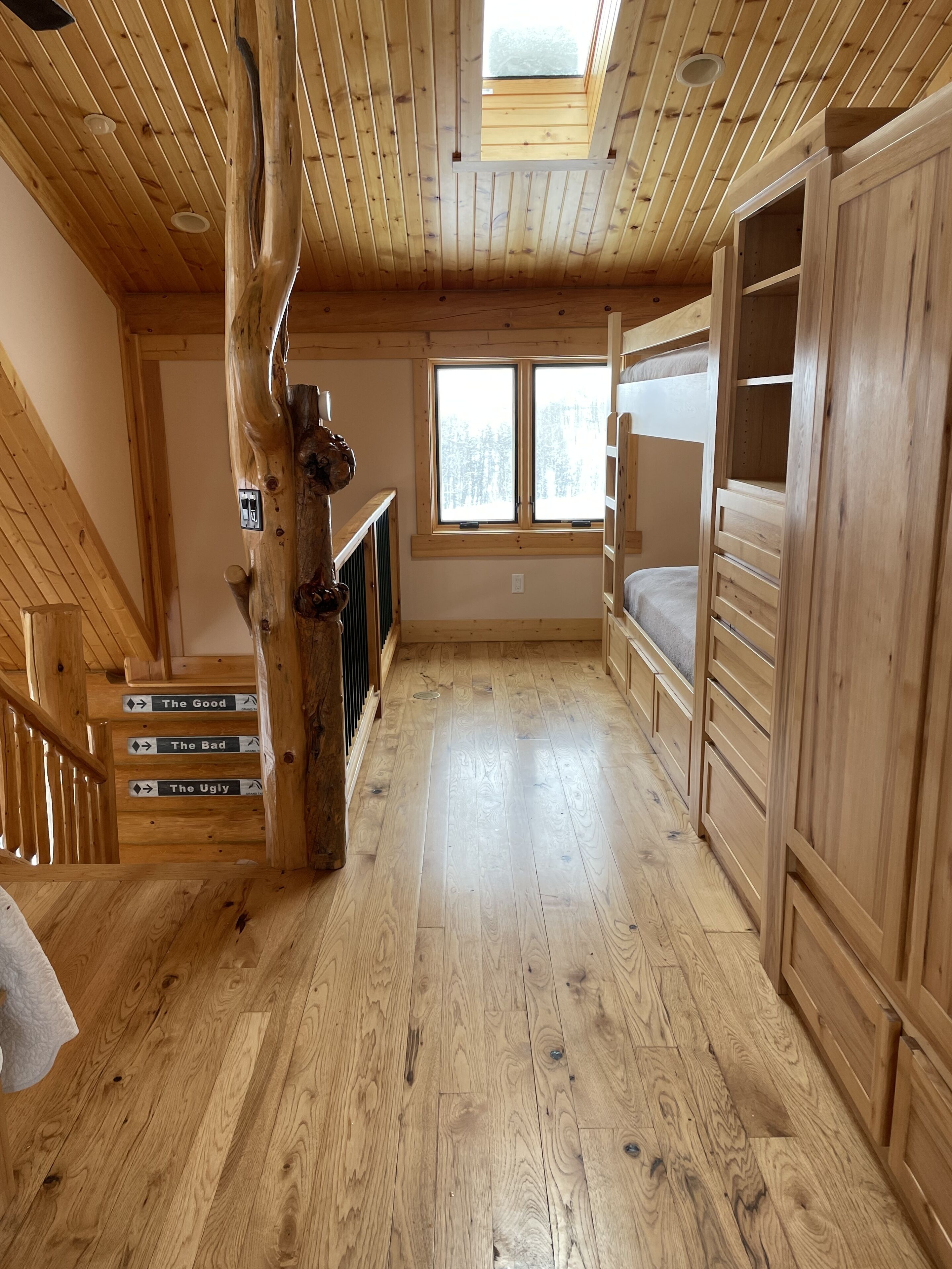 A wooden cabin interior with a loft bed, a staircase with directional signs, and a large window.