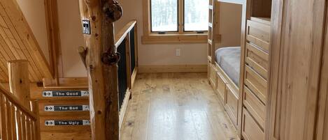 A wooden cabin interior with a loft bed, a staircase with directional signs, and a large window.