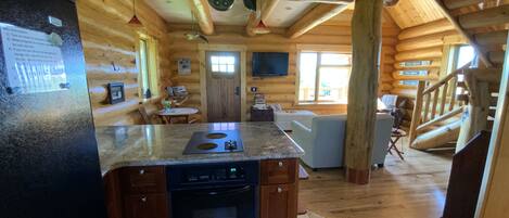 Interior d'una cabana de fusta amb cuina que inclou taulell de granat, rentavaixelles i forn integrat. Hi ha una zona de dinar amb taula i cadires, i una escala que mena a un altre nivell.