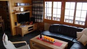 Een woonkamer met een zwarte leren bank, een houten salontafel met kaarsen, een televisie op een houten dressoir en een ruitjesgordijn.