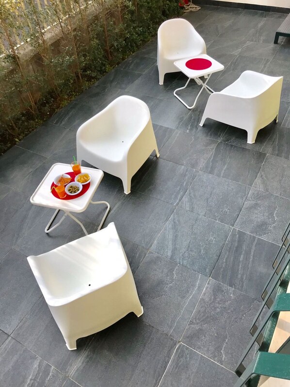 A patio area with four white plastic chairs and a small table, each with a red circular cushion. A tray with snacks and drinks is placed on the table.