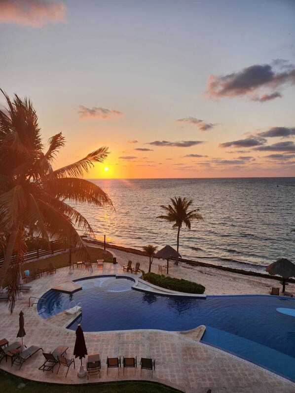 Une piscine d’un complexe hôtelier offrant une vue sur le coucher de soleil sur l’océan, entourée de palmiers et de chaises longues.
