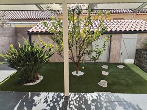Un patio con césped artificial, un árbol con flores amarillas, una planta grande y un techo de tejas al fondo.