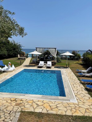 Una piscina con sillas de descanso y sombrillas, una terraza de piedra y vistas al mar.