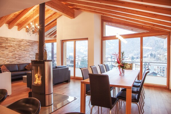 Ein geräumiges Wohnzimmer mit großem Holztisch, bequemen Sitzgelegenheiten, Kamin und großen Fenstern mit Blick auf die Berge.