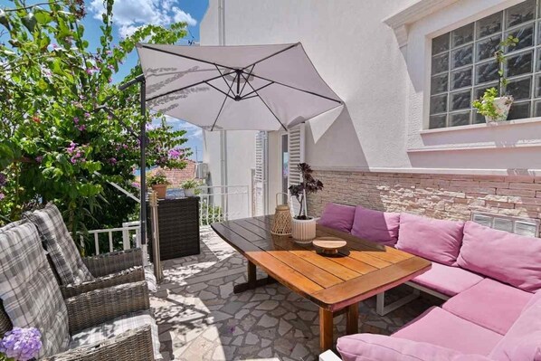 Une terrasse extérieure avec une table en bois et des meubles en osier aux couleurs rose et gris, un parasol blanc et une plante en pot placée sur un mur blanc.