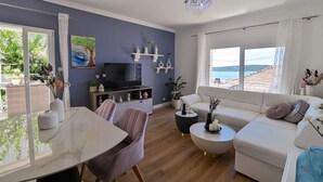 Ein modernes Wohnzimmer mit einem weißen Sofa, einem Esstisch und einem Fernseher. Sichtregale und eine Pendelleuchte sind ebenfalls vorhanden.