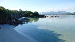 Ein Strand mit klarem Wasser, felsigem Ufer und Bergen im Hintergrund.