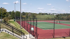 A tennis court with a red surface, surrounded by a chain-link fence, and a staircase leading up to a walkway.