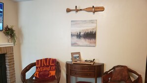 A cozy living room with a fireplace, two wooden chairs, a small side table, and a wall-mounted wooden paddle.