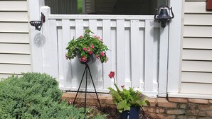A porch with white railings, a potted plant with pink flowers, and a small green shrub.