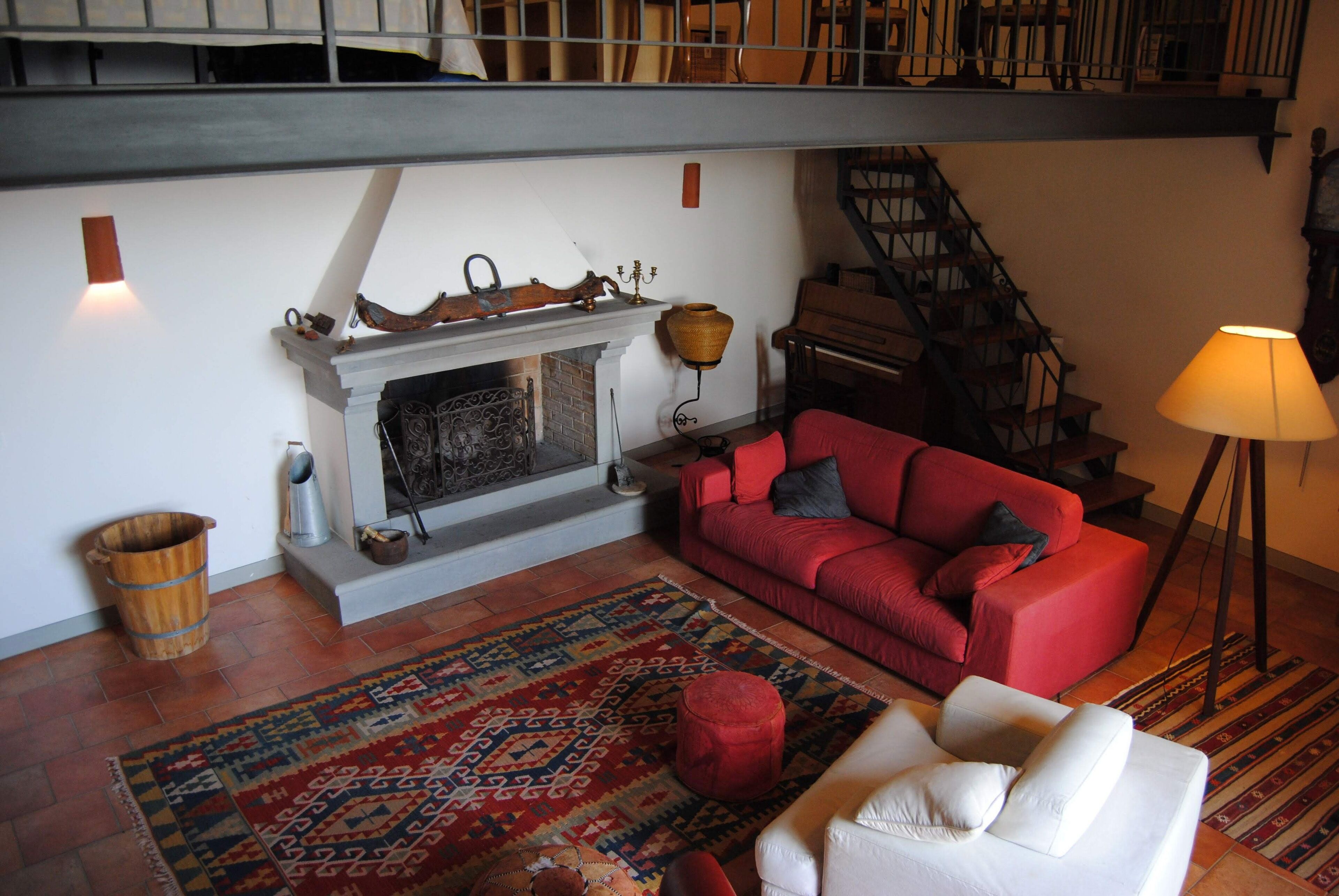 Ein Wohnzimmer mit Kamin, einem roten Sofa, einem weißen Sessel, einem gemusterten Teppich und einer Treppe.