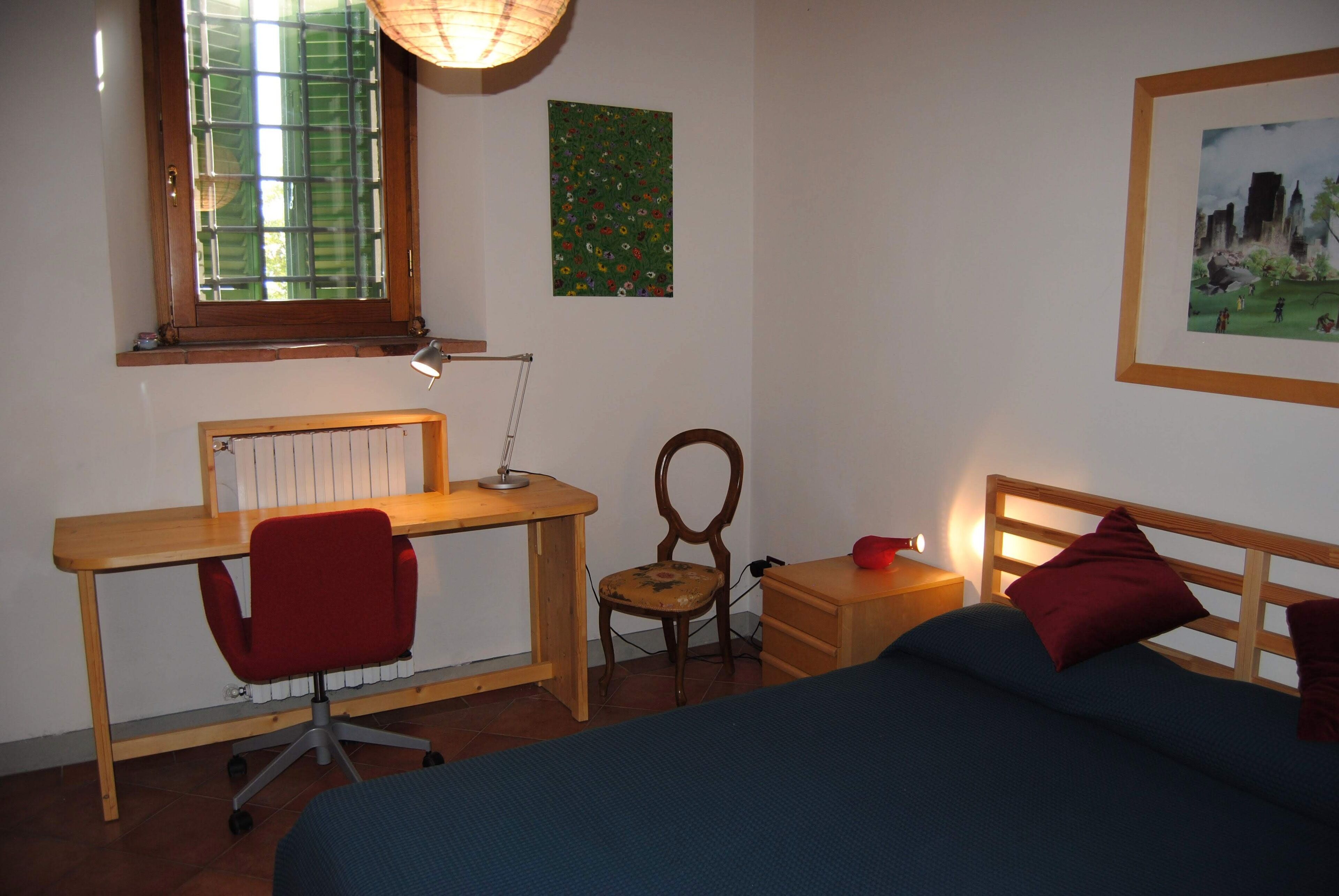 Une chambre à coucher avec un bureau en bois, une chaise de bureau rouge, un lit simple avec une tête de lit en bois, une table de chevet et une lampe.