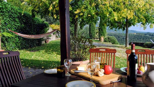 Une table de jardin dressée pour un repas en plein air, avec des assiettes, des verres, une bouteille de vin et une planche à découper sur laquelle se trouvent des aliments.
