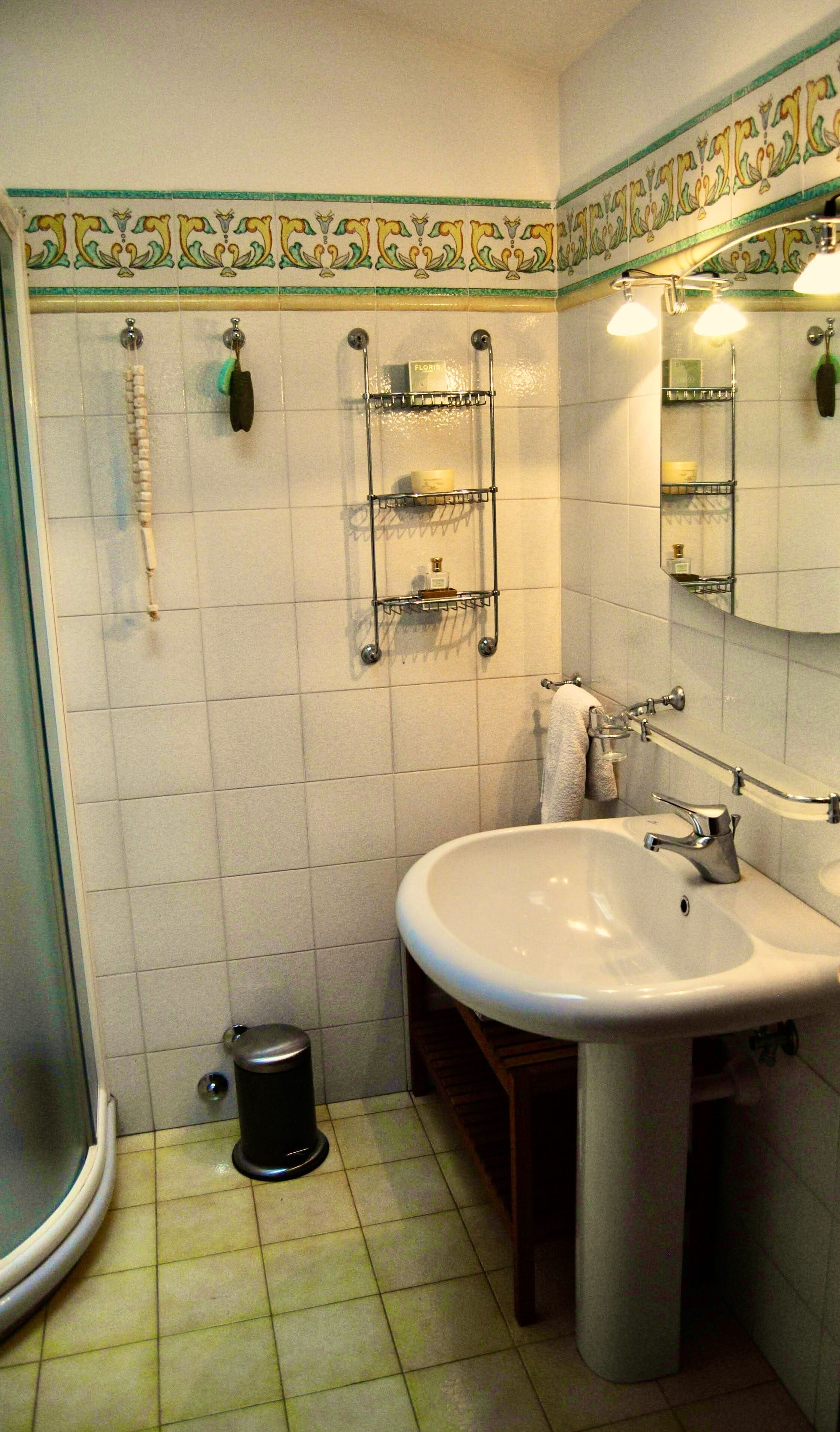 Un baño con lavamanos blanco, un espejo y ducha. Hay repisas con artículos de aseo y un cubo de basura.