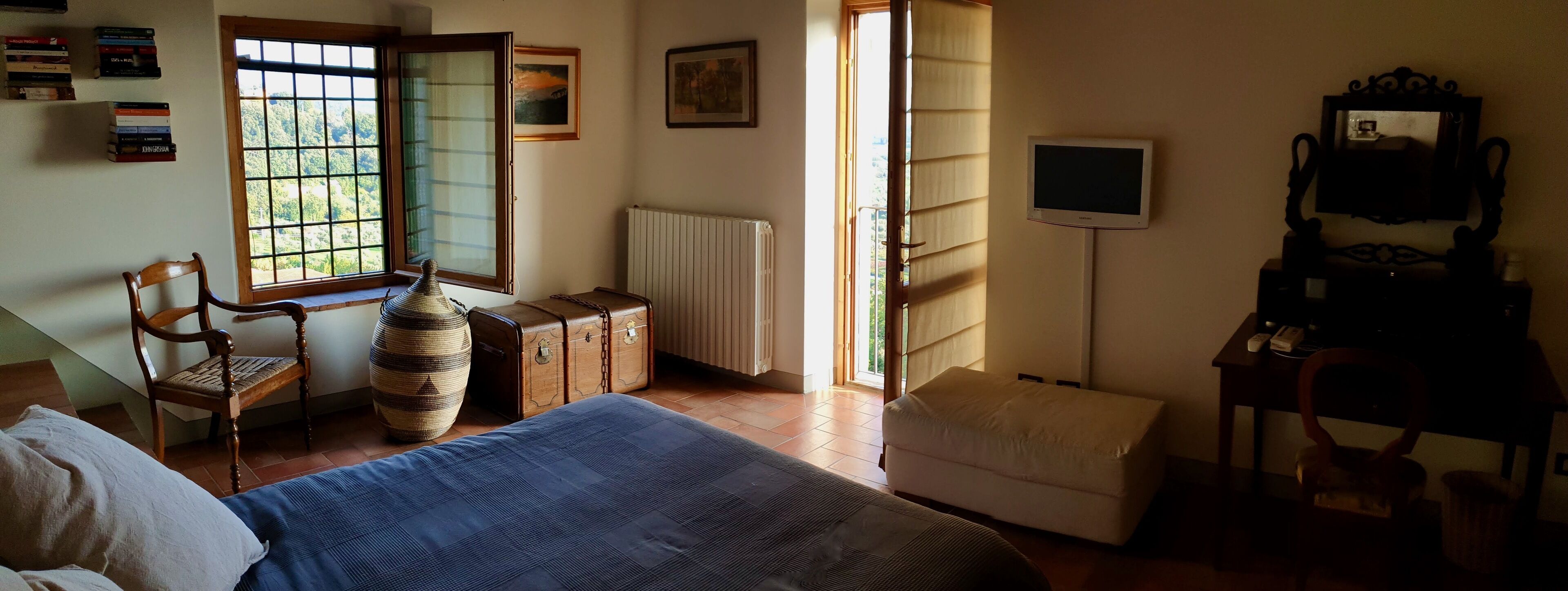 Ein Schlafzimmer mit Bett, Stuhl, Kommode, Heizkörper, Fernseher und einem Fenster mit Blick auf Grünflächen.
