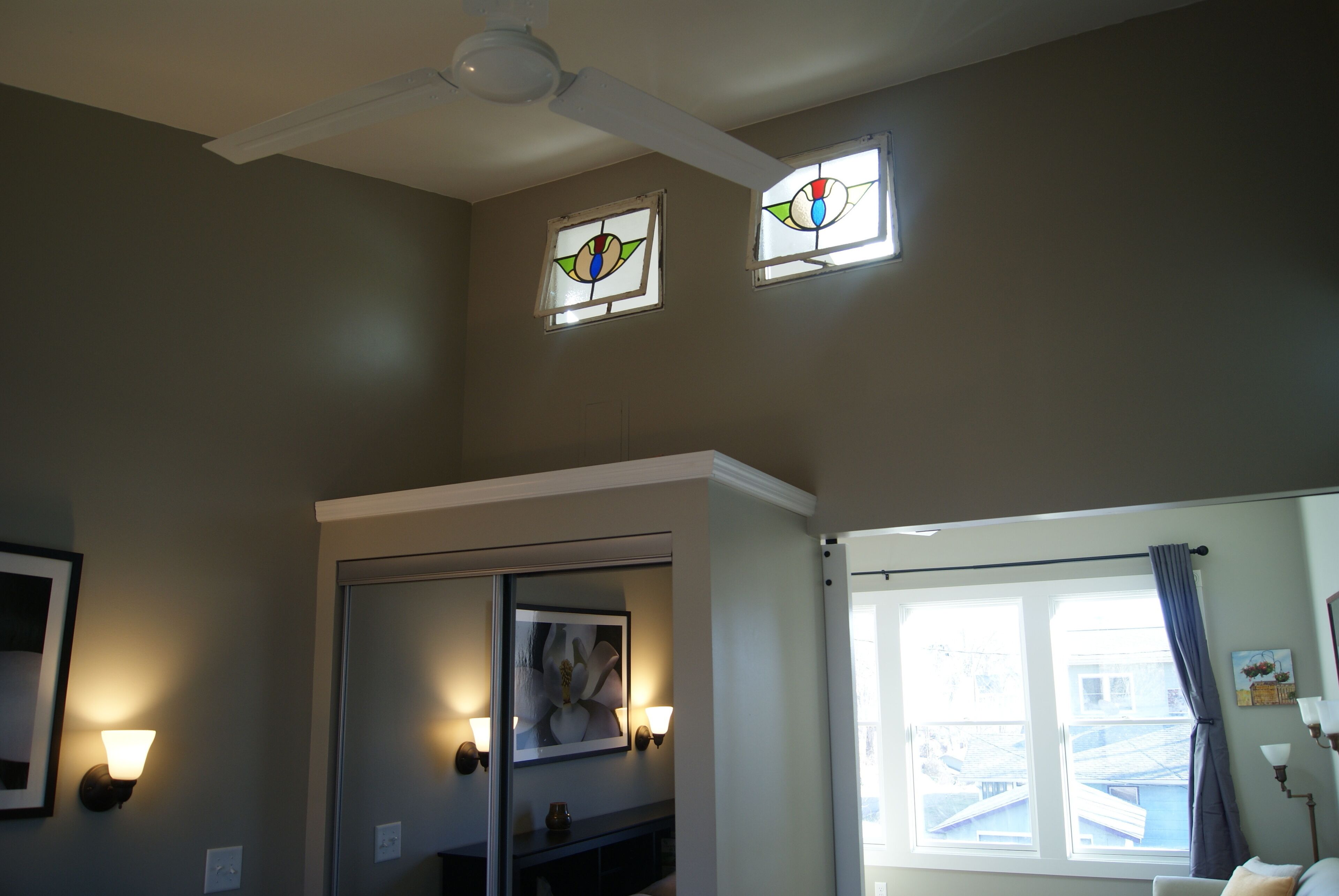 A room with a ceiling fan, two windows with stained glass, a framed artwork, and a door.