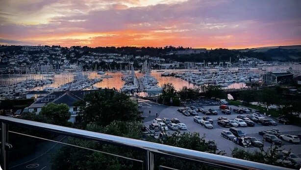 A marina with boats, a parking lot, and a town in the background during sunset. - The Loft: Amazing views, parking & fantastic location! (Kingswear)