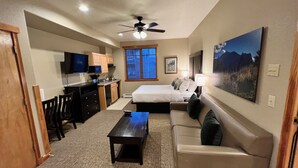 Ein kompaktes Hotelzimmer mit Bett, Sofa, kleinem Tisch, Fernseher, Deckenventilator, Fenster mit Jalousien und einem Bild an der Wand.