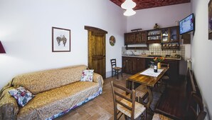 A cozy living room with a sofa, wooden dining table, and chairs. A kitchen with wooden cabinets and a tiled backsplash is visible in the background.