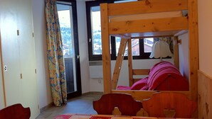 Een kamer met een stapelbed, een tafel met tafelkleed, een stoel, een raam met gordijnen en een deur.