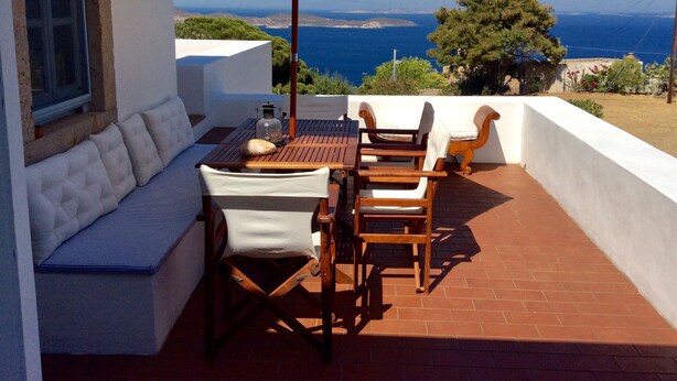 A terrace with a white cushioned bench, wooden chairs, a table, and an umbrella. A view of the sea and distant hills is visible. - Traditional villa in Hora with sea view to the Aegean  (ΠΑΤΜΟΣ)