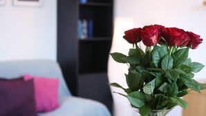 Quarto com cama, uma estante preta, um vaso com rosas vermelhas e uma mesa com revista e óculos de sol.