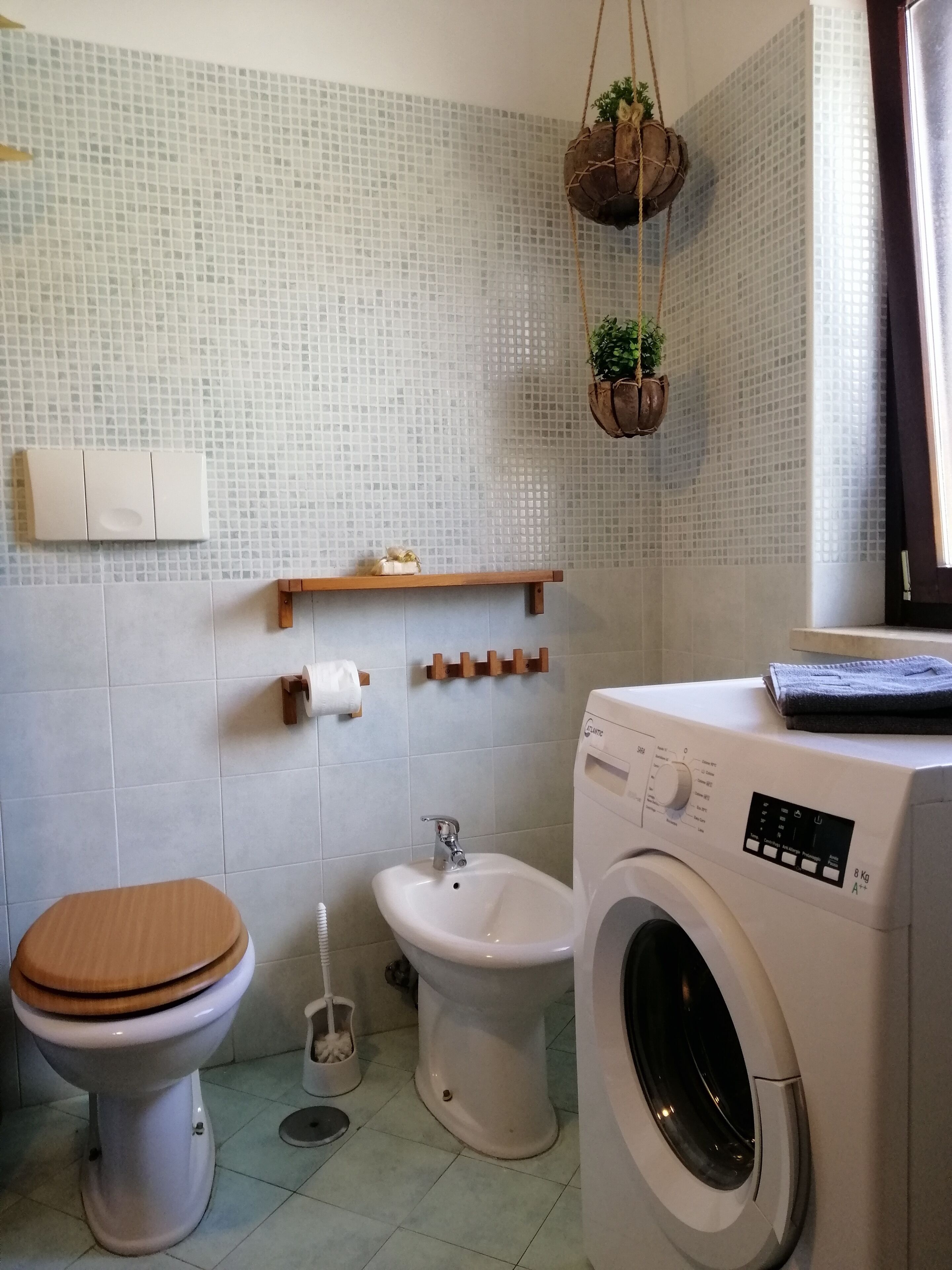 Ett badrum med toalett, bidé och tvättmaskin. Det finns hängande växter, en hylla med toalettartiklar och ett fönster med gardiner.