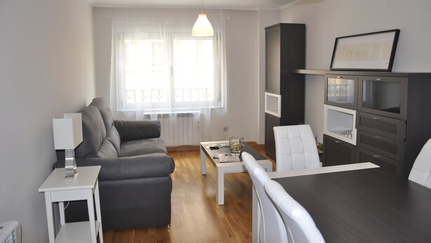 Una sala de estar moderna con un sofá gris, una mesa de centro blanca y una mesa de comedor con sillas. Hay un ventanal con cortinas translúcidas, una lámpara blanca y un cuadro en la pared.