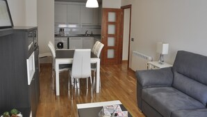 Una sala de estar moderna con un sofá gris, un comedor con mesa y sillas, una cocina con armarios blancos y una puerta de madera.