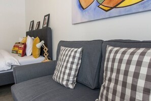 Ein modernes Wohnzimmer mit einer grauen Couch, karierten Kissen und einem kleinen Tisch mit Büchern und einer Lampe.