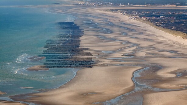 An aerial view of a coastal area with a long, narrow strip of land extending into the sea, surrounded by sand dunes and a clear sky.