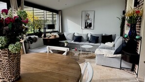 A modern living room with a dining table, grey sofas, a white armchair, a small round table, and a large window with curtains.